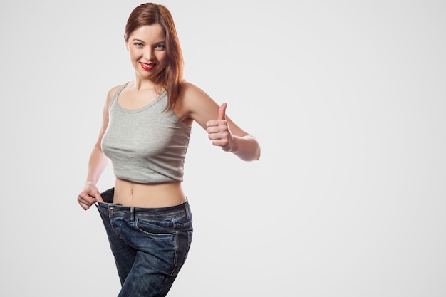 Photo portrait of happy beautiful slim waist of young woman standing in big jeans and gray top showing successful weight loss, indoor, studio shot, isolated on light gray background, diet concept.