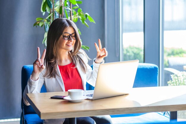 Portrait of happy beautiful stylish brunette young woman in glasses sitting, looking at her laptop monitor on video call with victory peace sign. indoor studio shot, cafe, office background.