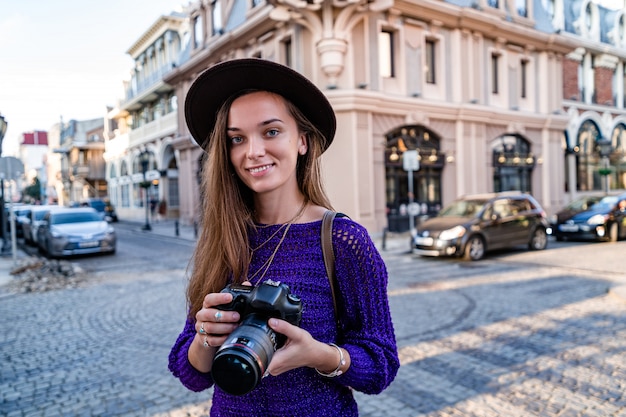 Photo portrait of happy beautiful stylish woman photographer in hat with a digital dslr camera taking photos while walking around the european city