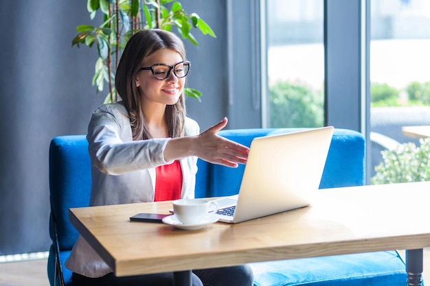 Portrait of happy beautiful stylish young woman in glasses sitting, looking at her laptop screen on video call and giving hand to handshake, toothy smile. indoor studio shot, cafe, office background.