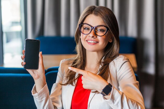 Portrait of happy beautiful stylish young woman in glasses sitting, showing and pointing at mobile smart phone screen and looking at camera with toothy smile. indoor studio shot, office background.