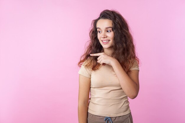 Portrait of happy beautiful teen girl with long curly brunette hair wearing casual style beige clothes pointing to the side and smiling, showing empty copy space. studio shot isolated, pink background