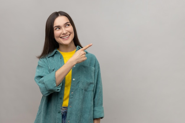 Portrait of happy beautiful young adult woman standing pointing and showing copyspace with happy face and toothy smile wearing casual style jacket Indoor studio shot isolated on gray background