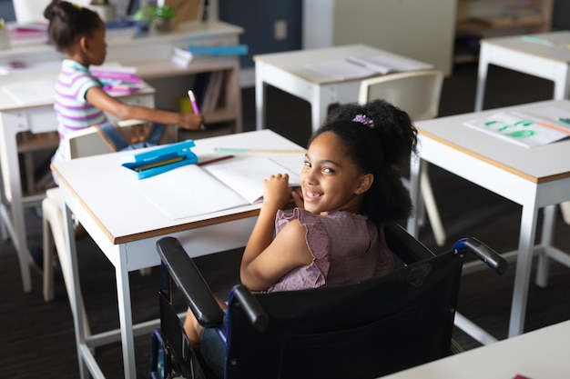 Portrait of happy biracial elementary schoolgirl sitting on wheelchair at desk in classroom