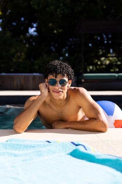 Portrait of happy biracial fit man in sunglasses standing in swimming pool in the sun, copy space. Summer, free time, relaxation and vacations.