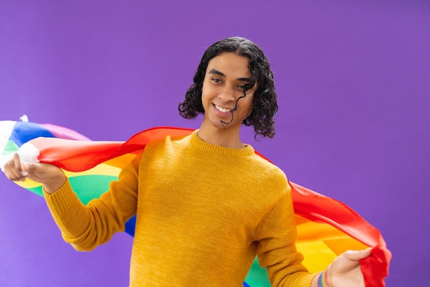 Photo portrait of happy biracial man holding lgbt rainbow flag and smiling on purple background. lgbt and pride concept.