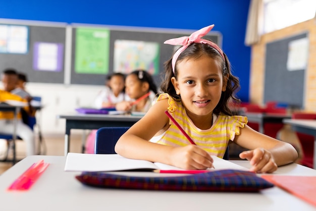 Portrait of happy biracial school girl sitting on desk in classroom studying at elementary school