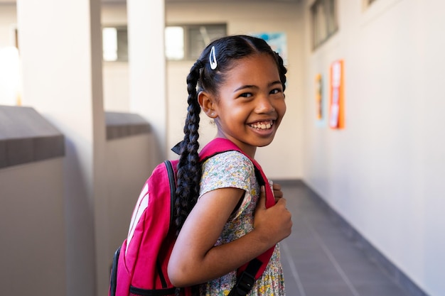 Photo portrait of happy biracial schoolgirl with school bag smiling in corridor at elementary school