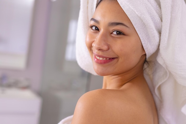 Portrait of happy biracial woman wearing towel smiling in bathroom, with copy space. Relaxation, self care, beauty, leisure and lifestyle concept.