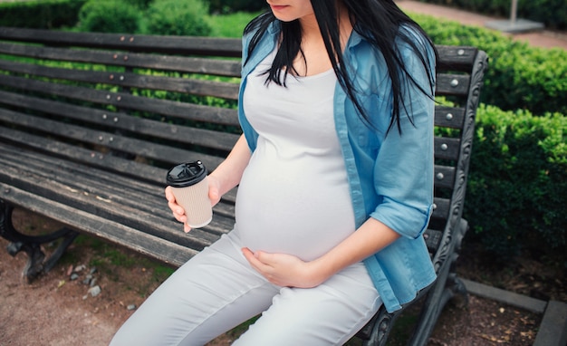 Portrait of a happy black hair and proud pregnant woman in park. The female model is sitting on a city bench and drinking coffee or tea.