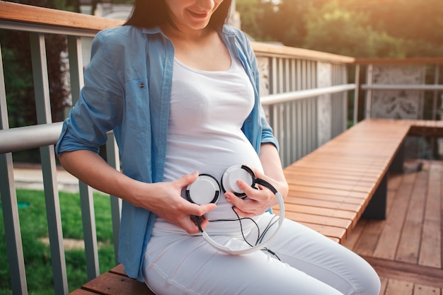 Ritratto di un felice capelli neri e orgogliosa donna incinta nel parco. è seduta su una panchina della città. la donna incinta sta ascoltando musica nel parco con un bambino non ancora nato