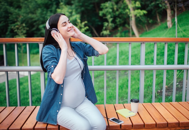 幸せな黒髪と公園で誇りに思っている妊娠中の女性の肖像画。彼女は街のベンチに座っています。妊娠中の母親は、胎児と公園で音楽を聴いています