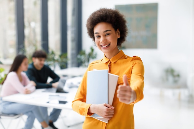 Photo portrait of happy black lady student showing thumb up standing in classroom with classmates studying