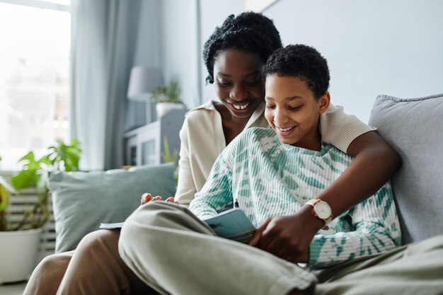 Portrait of happy black mother with daughter doing homework together while sitting on sofa at home