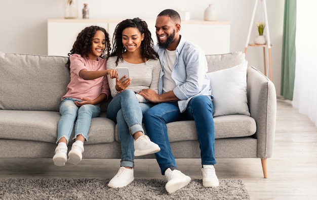 Portrait of happy black woman, man and girl holding and using smartphone sitting on the sofa in living room. People spending time together, watching video, choosing gifts, daughter pointing at gadget