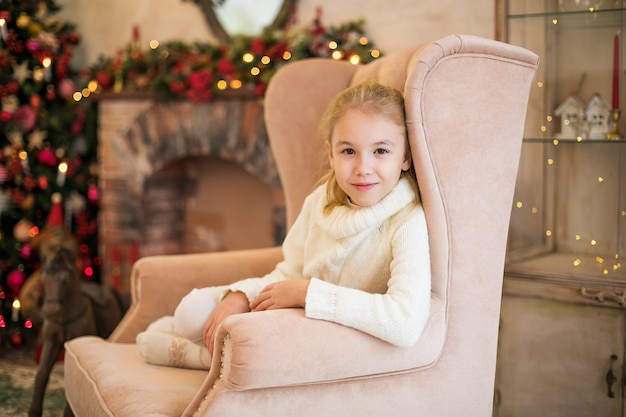  portrait of happy blonde child girl in white sweater siting on the floor near the Christmas tree