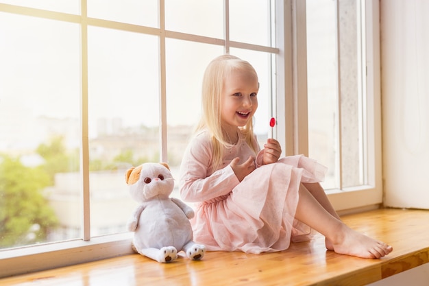 Portrait of happy blonde girl wearing pink dress holding a lollipop while sitting with a toy on a window sill