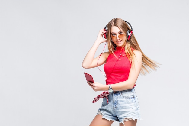 Portrait of happy blonde girl at white wall listens to music in headphones and looking to the front with smart phone.