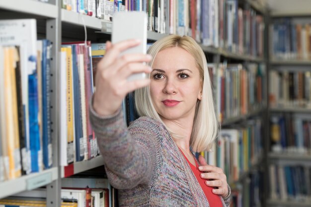 Портрет счастливой блондинки в библиотеке университета и делающей селфи с мобильным телефоном