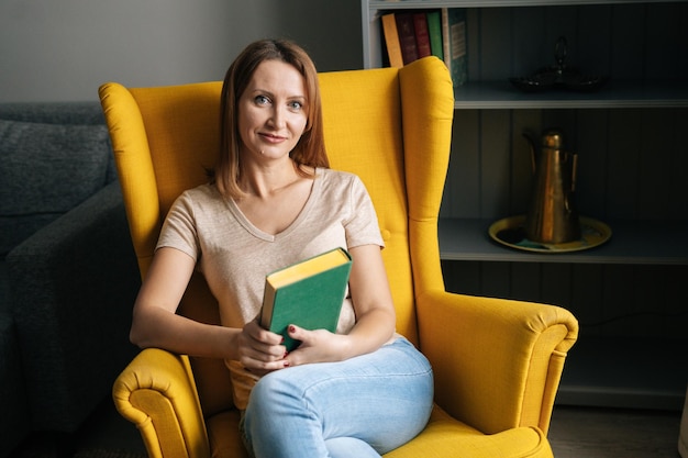 Portrait of happy blonde woman sitting in comfy yellow chair holding paper book in dark room with modern interior smiling looking at camera