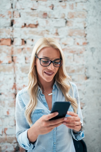 Portrait of happy blonde woman smiling against brick wall and chatting on mobile phone. 