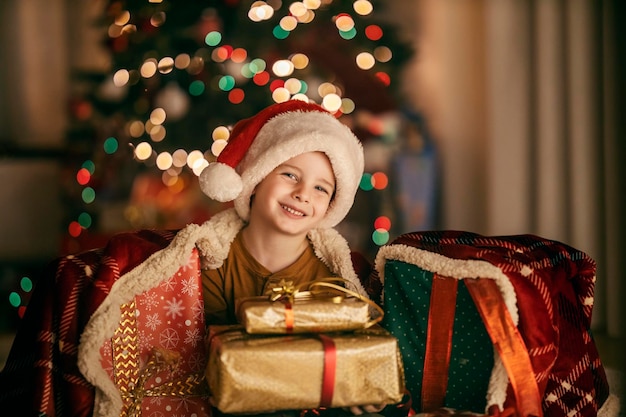 Портрет счастливого мальчика, сидящего дома в канун Рождества и Нового года, окруженного подарками
