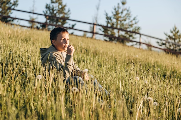 Portrait of happy boy sitting on top of hill in grass field and enjoying beautiful landscape at sunset Teenager hiker resting in nature Active lifestyle Concept of local travel