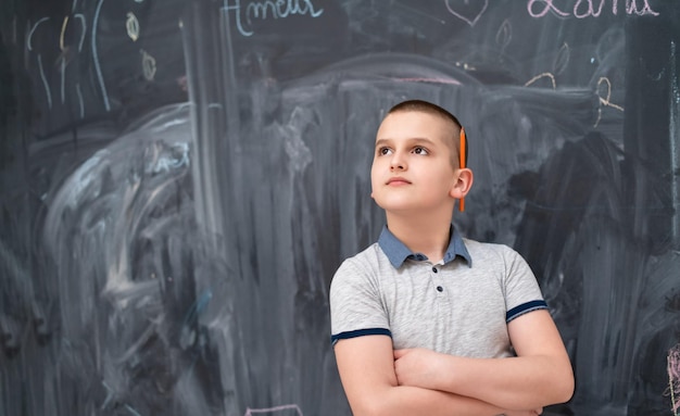 portrait of happy boy with orange wooden pen behind the ear standing in front of black chalkboard