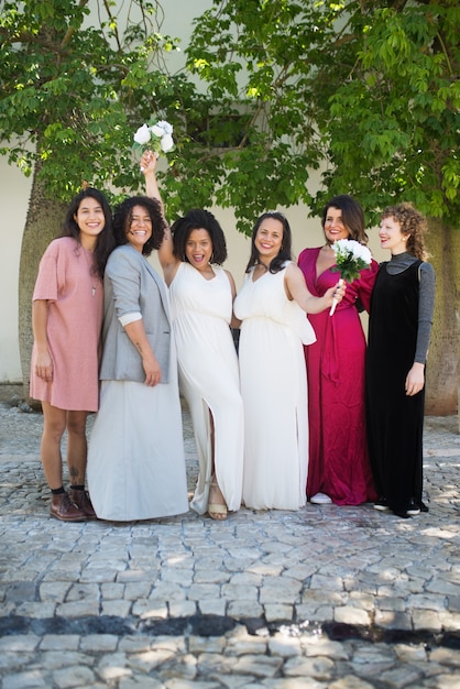 Portrait of happy brides and guests at wedding. Women of different nationalities in festive dresses standing together. Brides showing bouquets