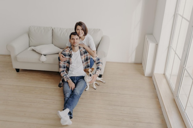 Portrait of happy brunette young woman embraces with love her husband who sits on floor with pedigree dog, pose in empty spacious room, move in new apartment after marriage, start family life