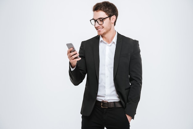 Portrait of a happy businessman in eyeglasses using smartphone over white wall