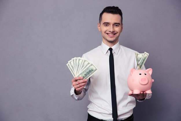 Portrait of a happy businessman holding pig money box and money over gray wall