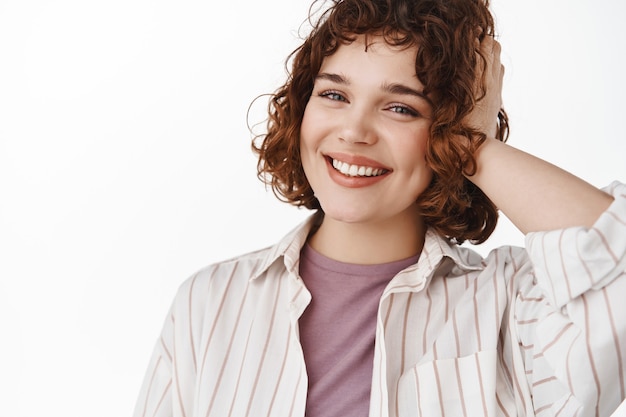 Portrait of happy candid girl with relaxed pose, cheerful white smile and stylish curly short hairstyle, touching hair and looking cheerful at cameraon white