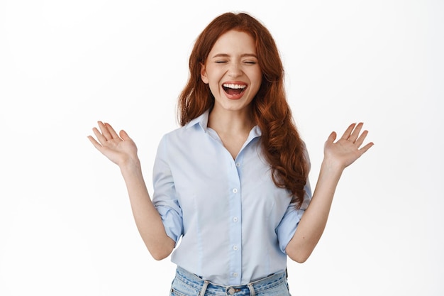 Portrait of happy candid redhead girl laughing, smiling with white teeth, spread hands sideways carefree and chuckle from funny joke, standing against white background