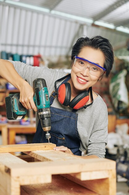 Portrait of happy carpenter in protective glasses using drill when making wooden furniture in workshop