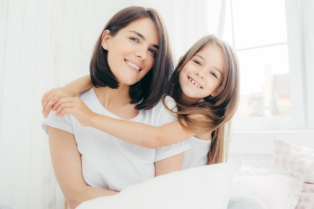 Portrait of happy Caucasian mother with charming smile and her small daughter embraces with love mum being in good mood pose on comfortable bed against cozy interior People and family concept