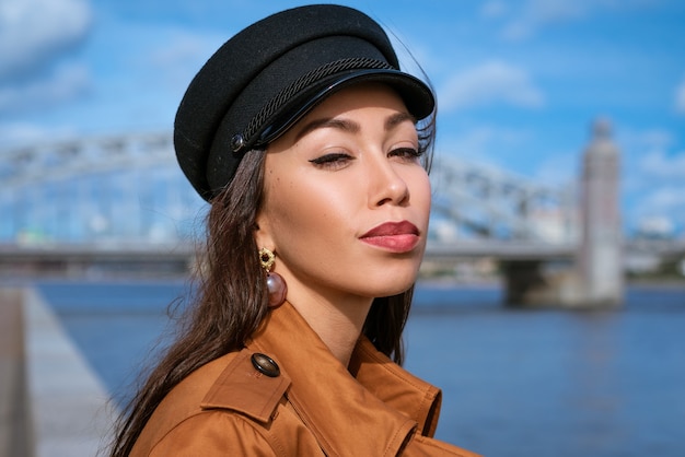 Portrait of a happy caucasian woman on the embankment of the river in a black cap and brown jacket o...