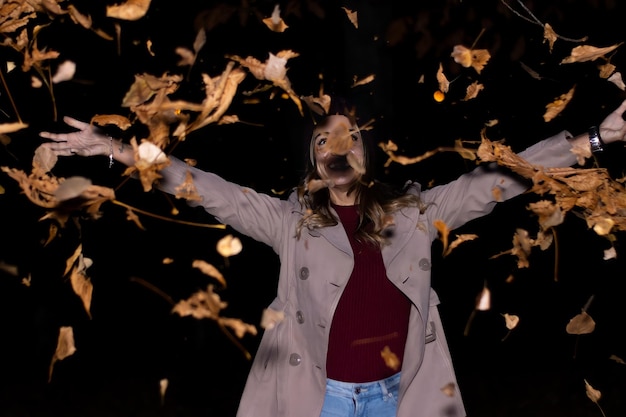 Portrait of happy caucasian woman throwing leaves at night in autumn