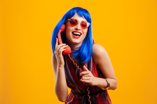 Portrait of happy caucasian woman wearing blue wig and sunglasses holding handset and laughing isolated over yellow wall