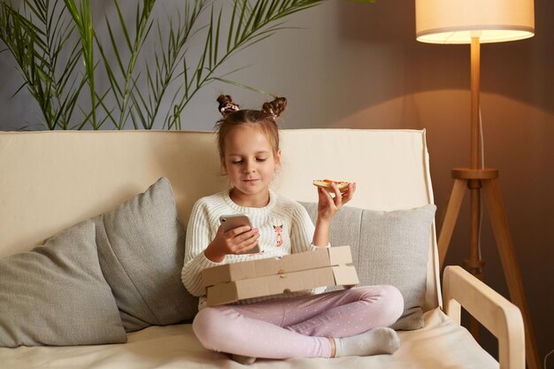 Portrait of happy charming cute little girl sitting on cough with crossed legs and eating pizza having snack while using mobile phone watching cartoons while having dinner