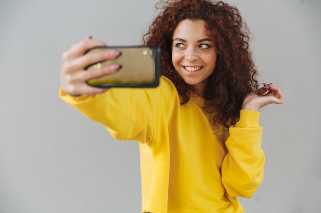 Portrait of happy cheerful beautiful curly woman isolated over gray wall using mobile phone take a selfie.