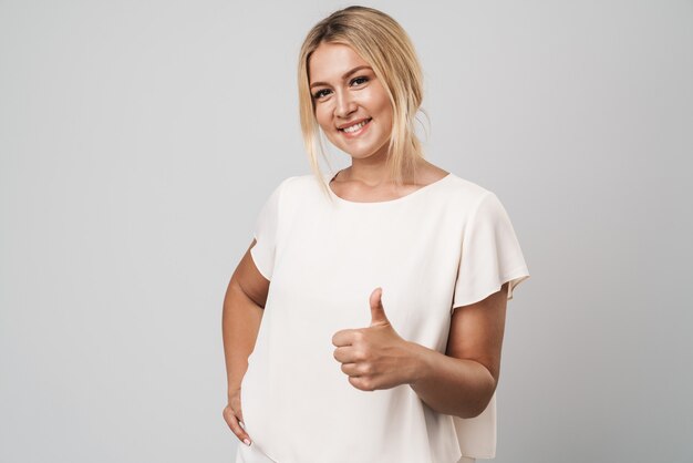 Portrait of a happy cheerful beautiful young amazing blonde woman posing isolated over grey wall dressed in basic white t-shirt showing thumbs up gesture