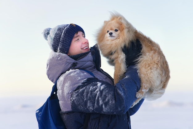 Portrait of happy cheerful positive guy, handsome man is having fun, playing in snow with his cute little puppy Pomeranian Spitz dog at winter snowy day in warm clothes, smiling. People love animals