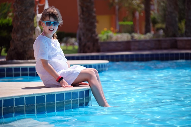 Портрет счастливой девочки в белом платье, отдыхающей у бассейна в солнечный летний день во время тропических каникул