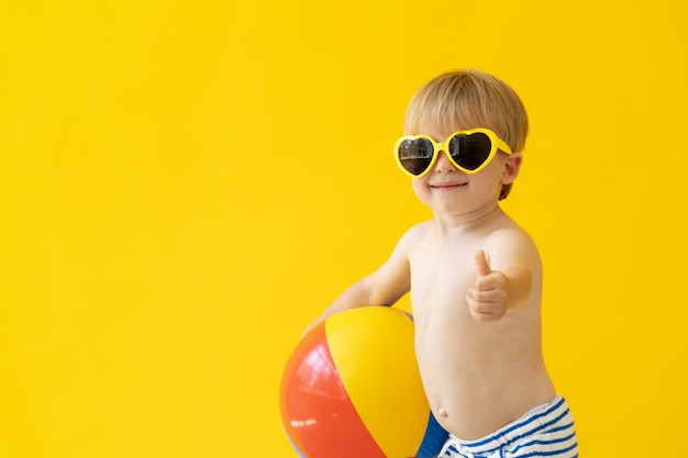 Portrait of happy child holding beach ball against yellow wall on summer vacation.