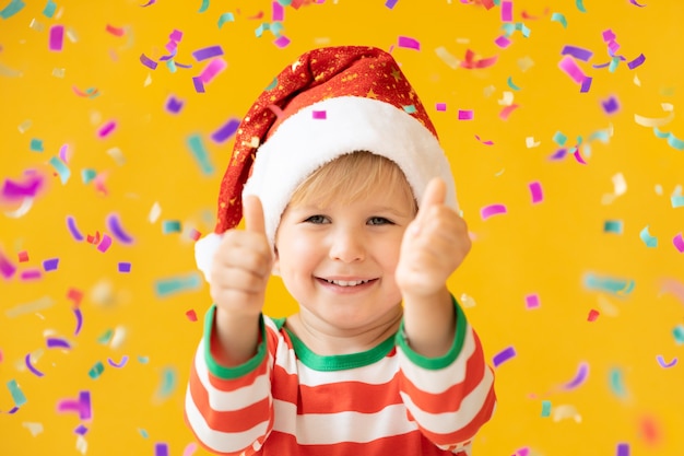 Photo portrait of happy child showing thumbs up. funny kid dressed santa claus hat against yellow background. christmas holiday concept