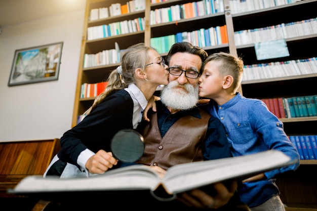 Ritratto di bambini felici, ragazzo e ragazza, baciando il loro vecchio nonno barbuto sulle guance mentre trascorrono del tempo, leggendo insieme libri straordinari in biblioteca o in un'accogliente stanza a casa