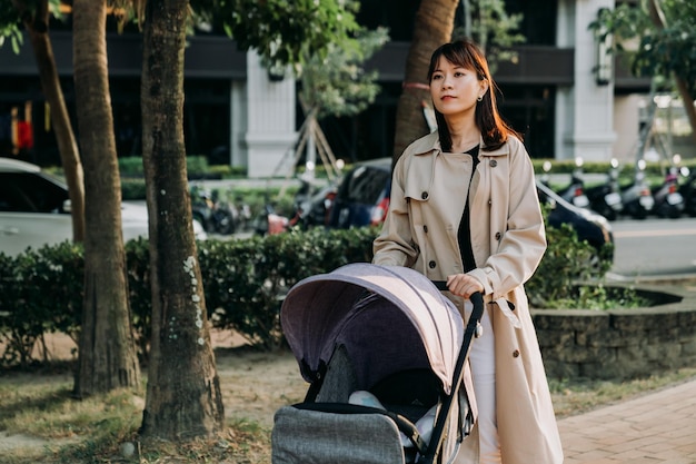 portrait happy chinese career mom is pushing walking with a baby pram at leisure in a park at urban area on an autumn sunny day.