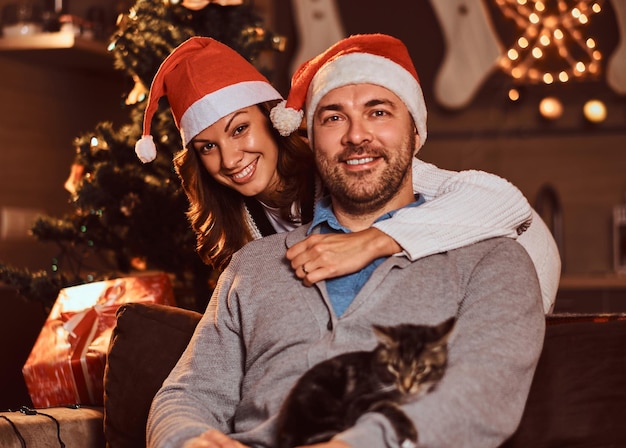 幸せなカップルの肖像画-魅力的な女性が男性を抱き締め、ラップトップを使用しています。クリスマスイブを祝う猫と女性。