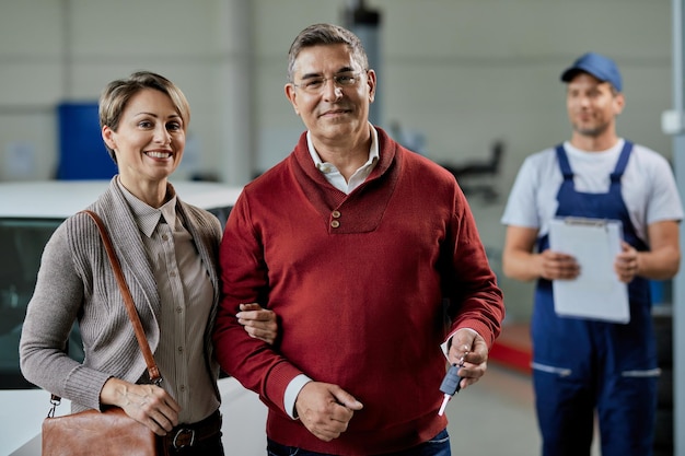 Portrait of happy couple holding key of their repaired car in a workshop Their mechanic in standing in the background